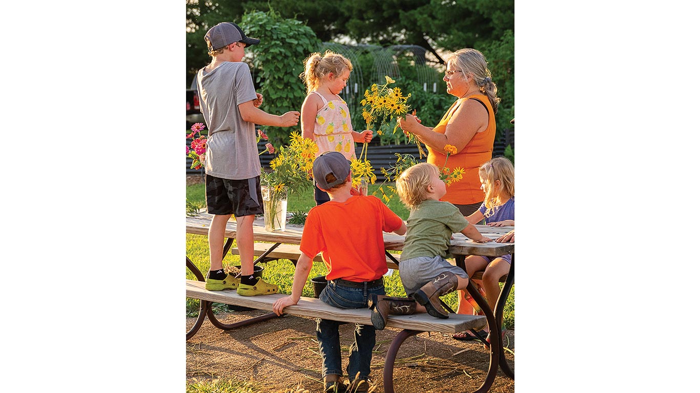 Group of kids and an adult arrange yellow flowers on a picnic table outdoors.