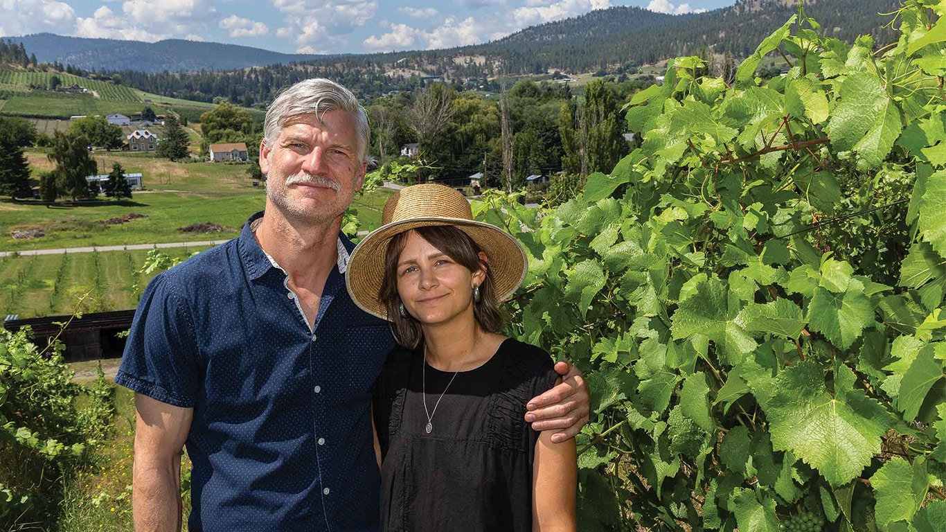 Two people stand beside grapevines in a hillside vineyard with rolling fields and mountains behind.
