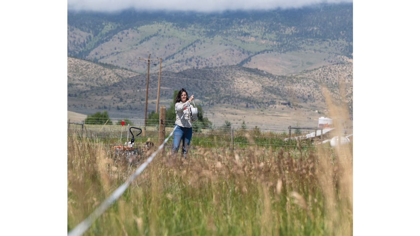 Person standing in a grassy field measuring a long tape line with mountains in the background.