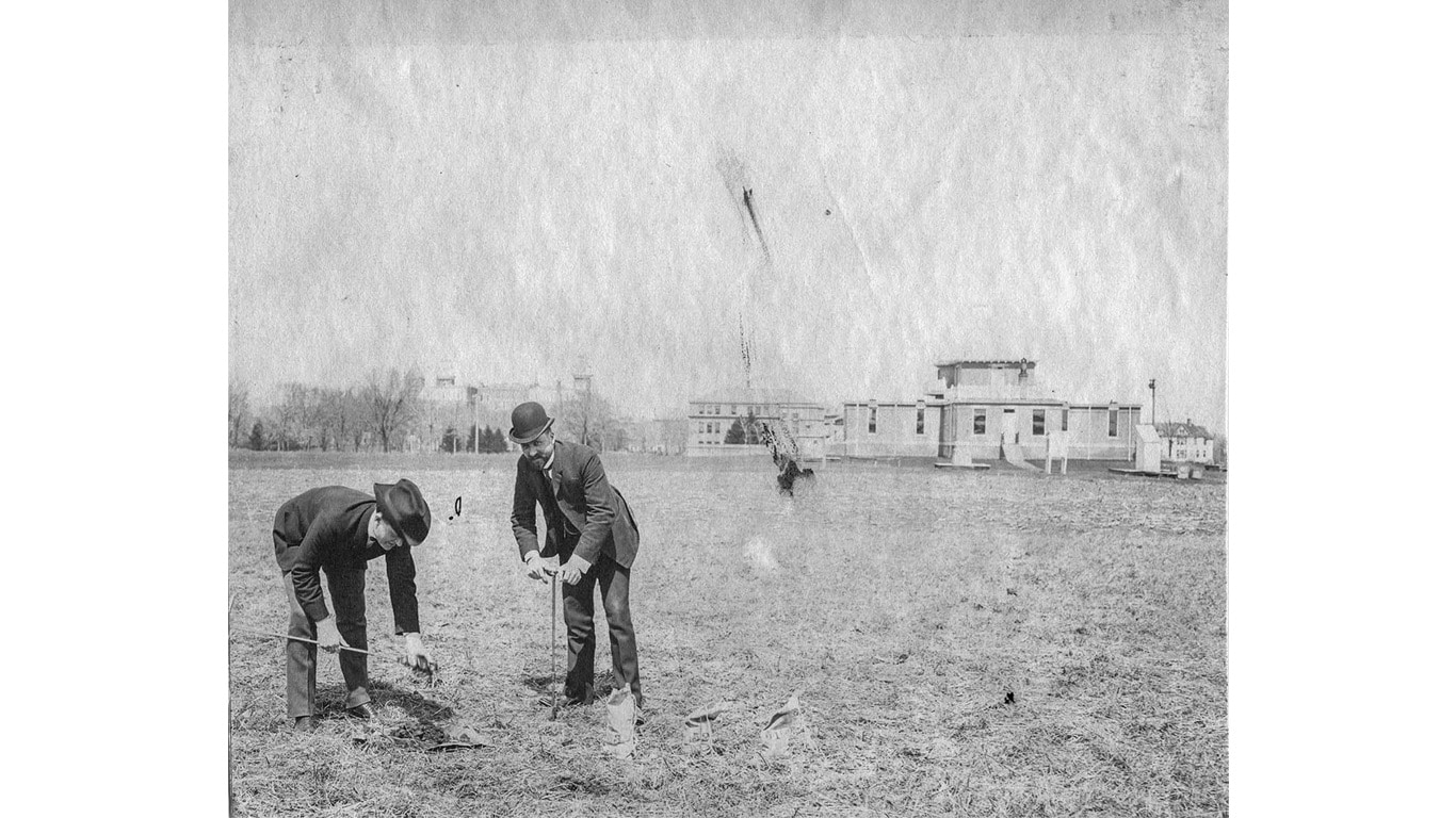 Two people take soil samples in an open field with early campus buildings visible in the distance.