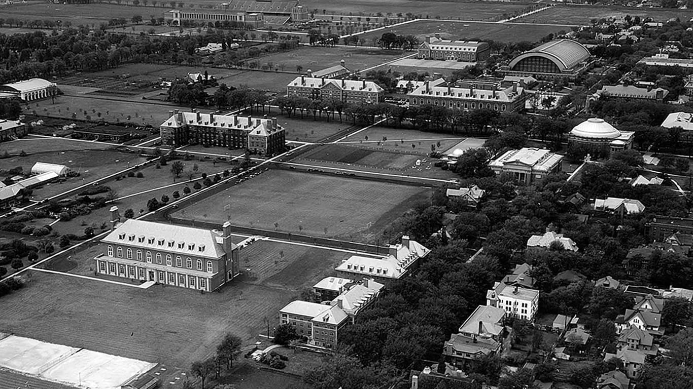 An aerial view of a university campus showing agricultural plots, academic buildings, and surrounding tree-lined areas.