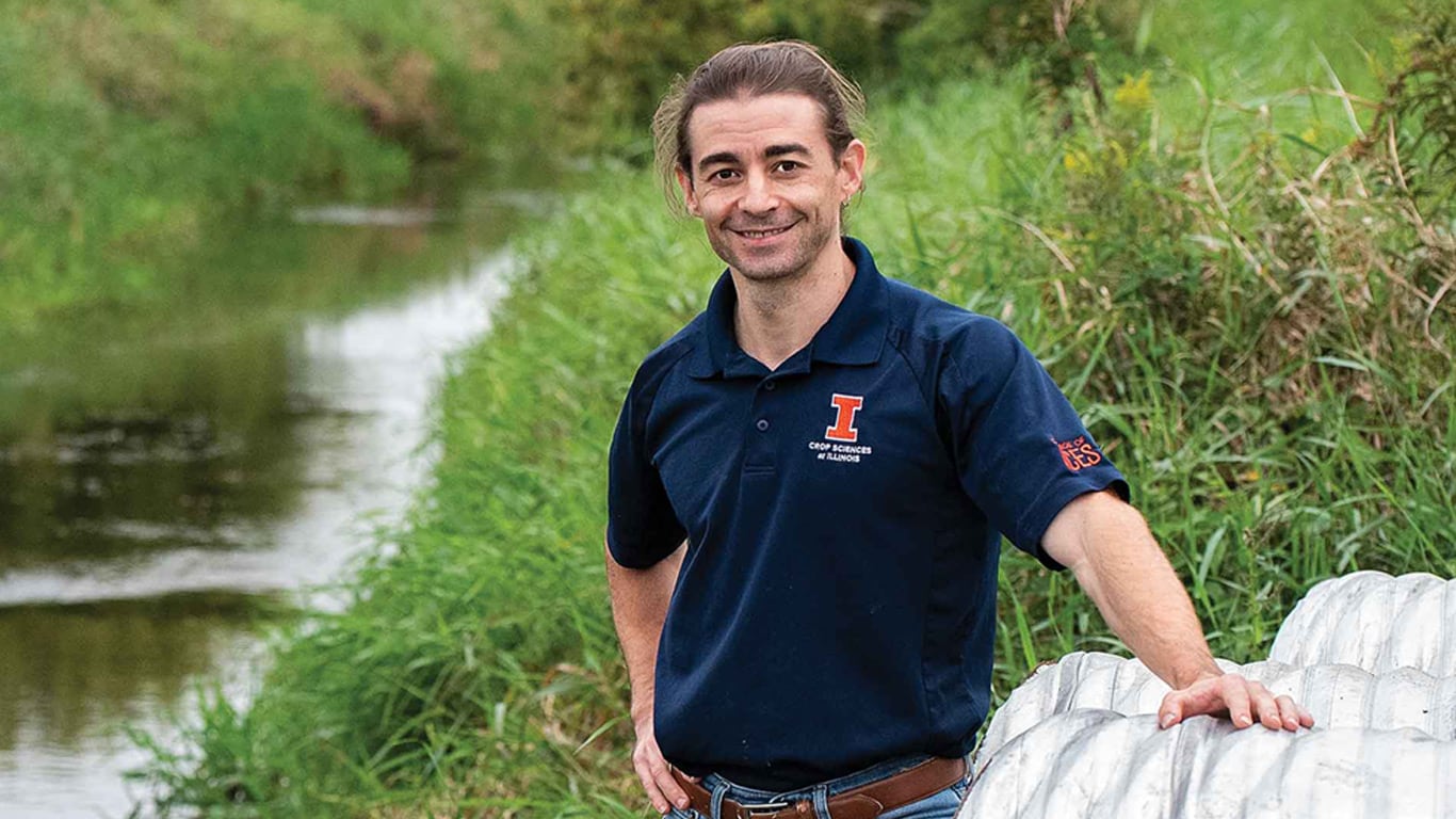 A person stands beside a metal drainage structure near a grassy waterway.