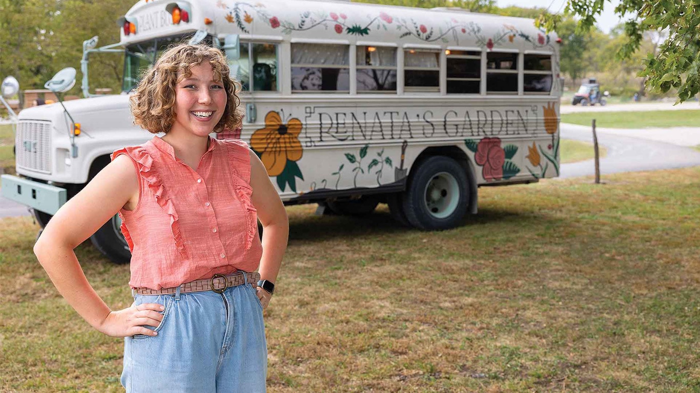 Person standing with hands on hips in front of a floral-painted short bus labeled 'Renata&rsquo;s Garden'.