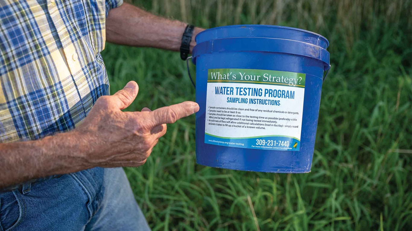 Hand points to blue bucket labeled &ldquo;Water Testing Program&rdquo; held over tall grass outdoors.
