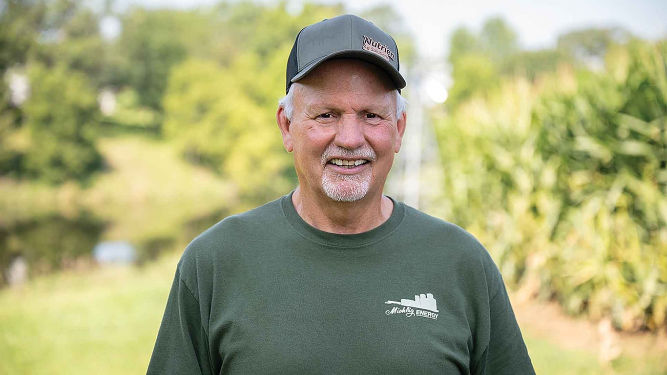 Person wearing a green 'Midwest Energy' shirt and cap stands outdoors near a cornfield and grassy path.