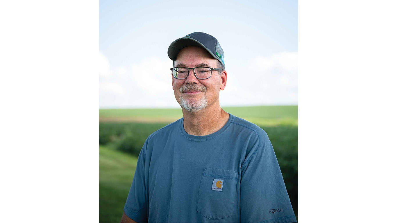 Person in a blue t-shirt and cap stands outdoors with rolling green fields and trees in the background.