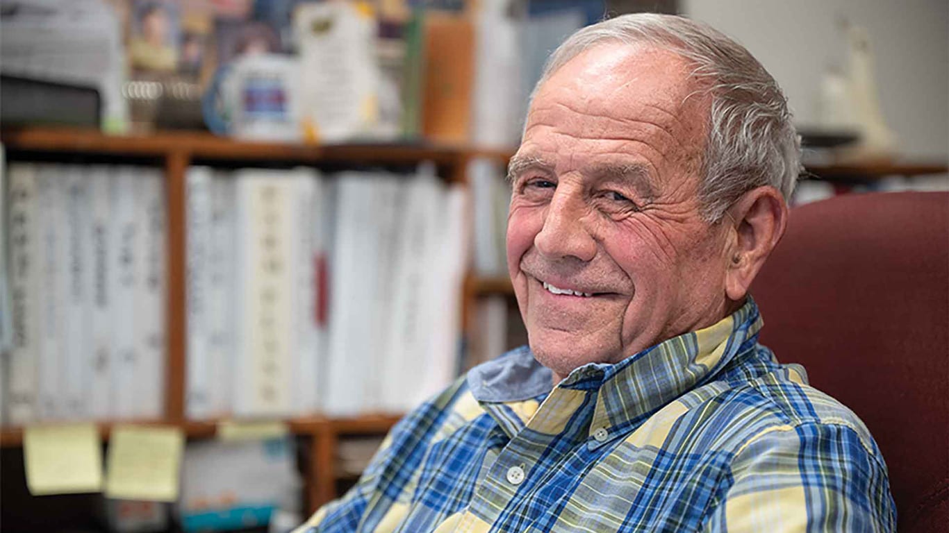 Person in a plaid shirt sits in an office chair with shelves of binders and paperwork in the background.