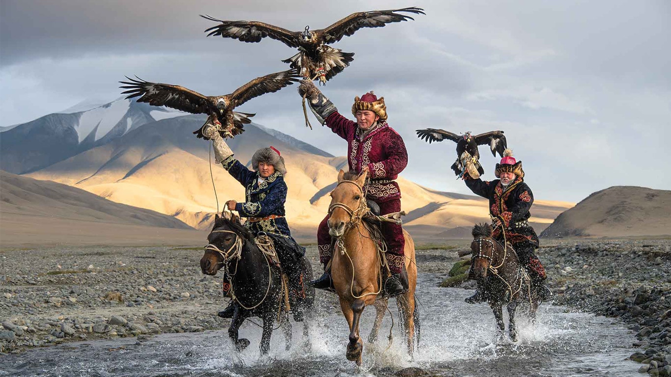 Three riders on horseback holding eagles while crossing a shallow stream with mountains behind them.