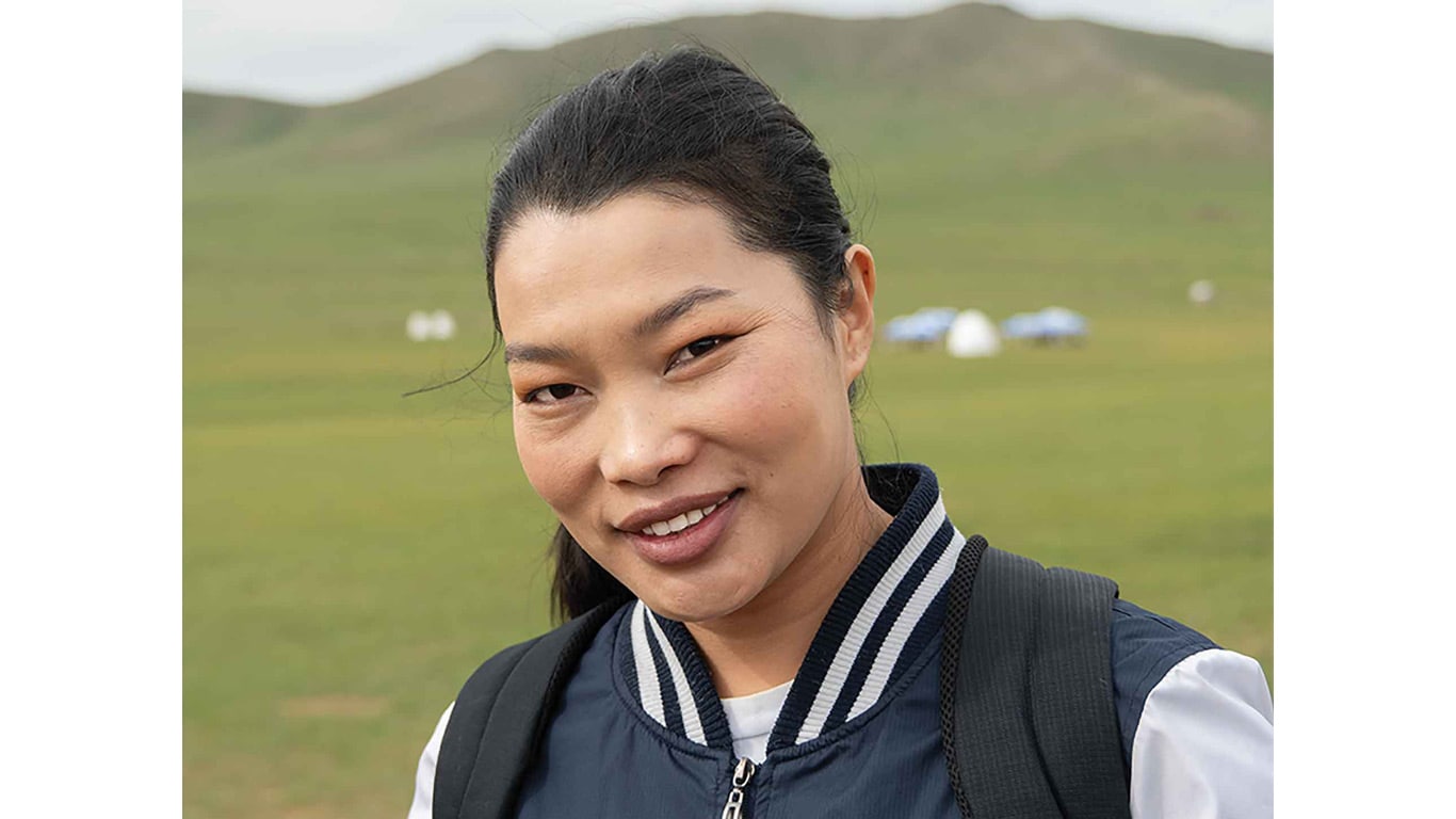 Person standing outdoors with grassy hills and tents in the background.