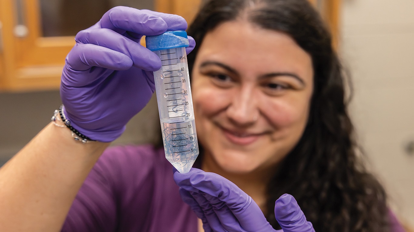 Gloved hands hold a graduated tube containing blue filaments in a lab setting.
