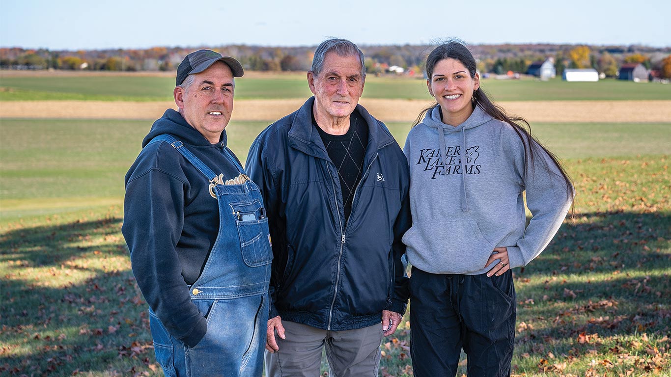 Three people standing together in a farm field with buildings in the background.