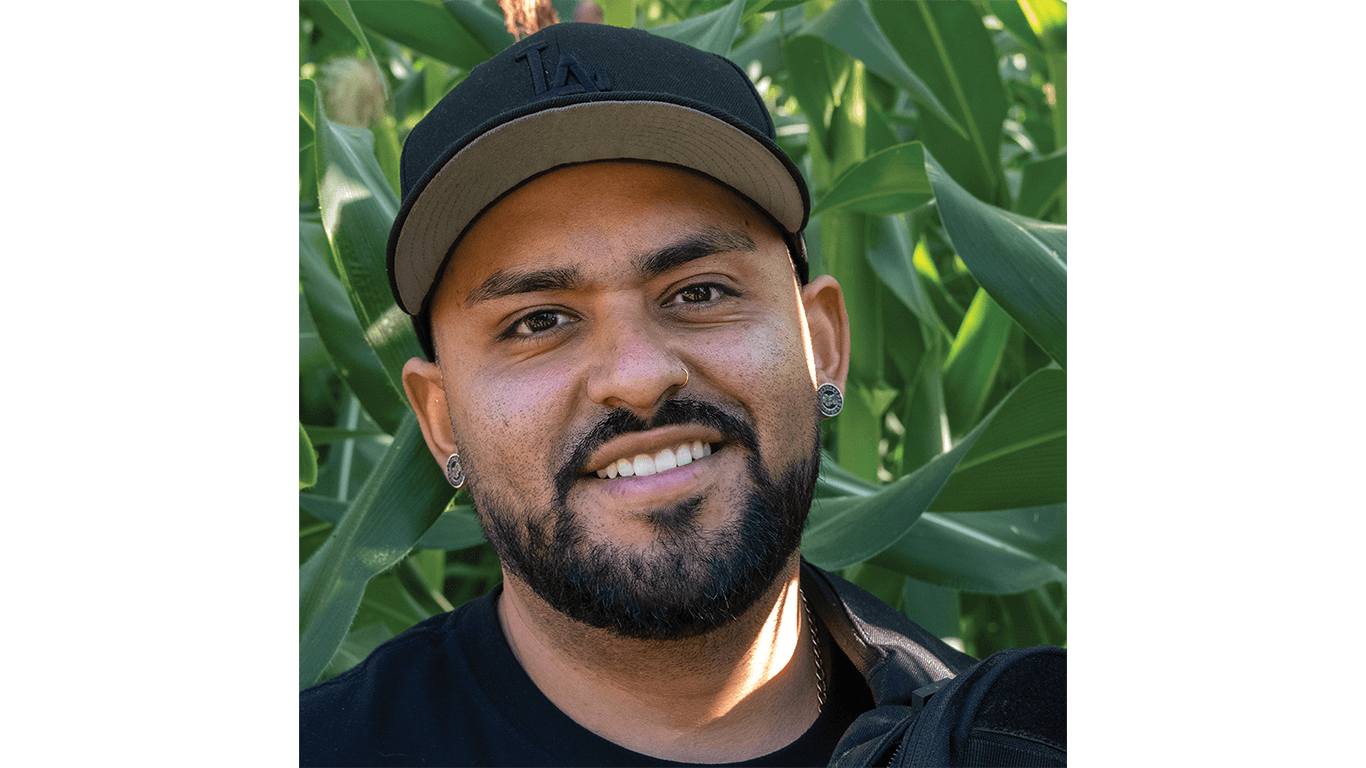 Person wearing black cap stands among tall green corn leaves, facing camera.