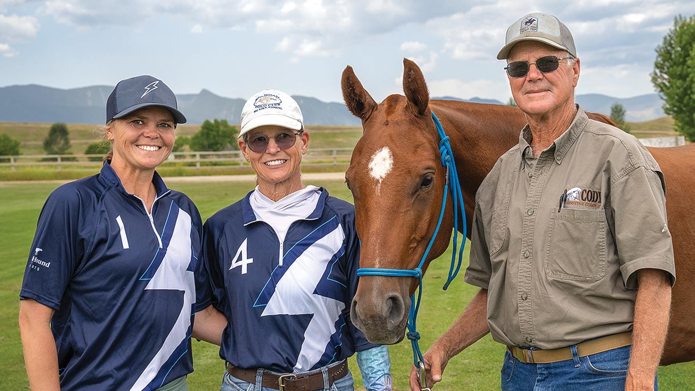 Three people standing beside a brown horse on a grassy field, with mountains in the distance.