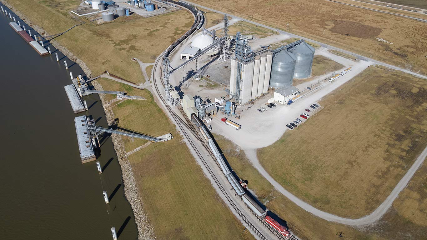Aerial view of grain facility with silos, rail cars, and river barge loading dock.