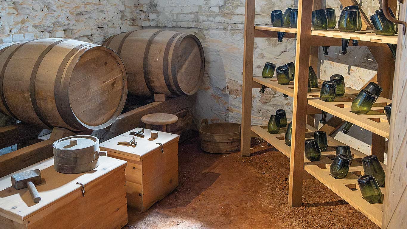 Wooden barrels and tools in a rustic interior with racks of inverted glass bottles.