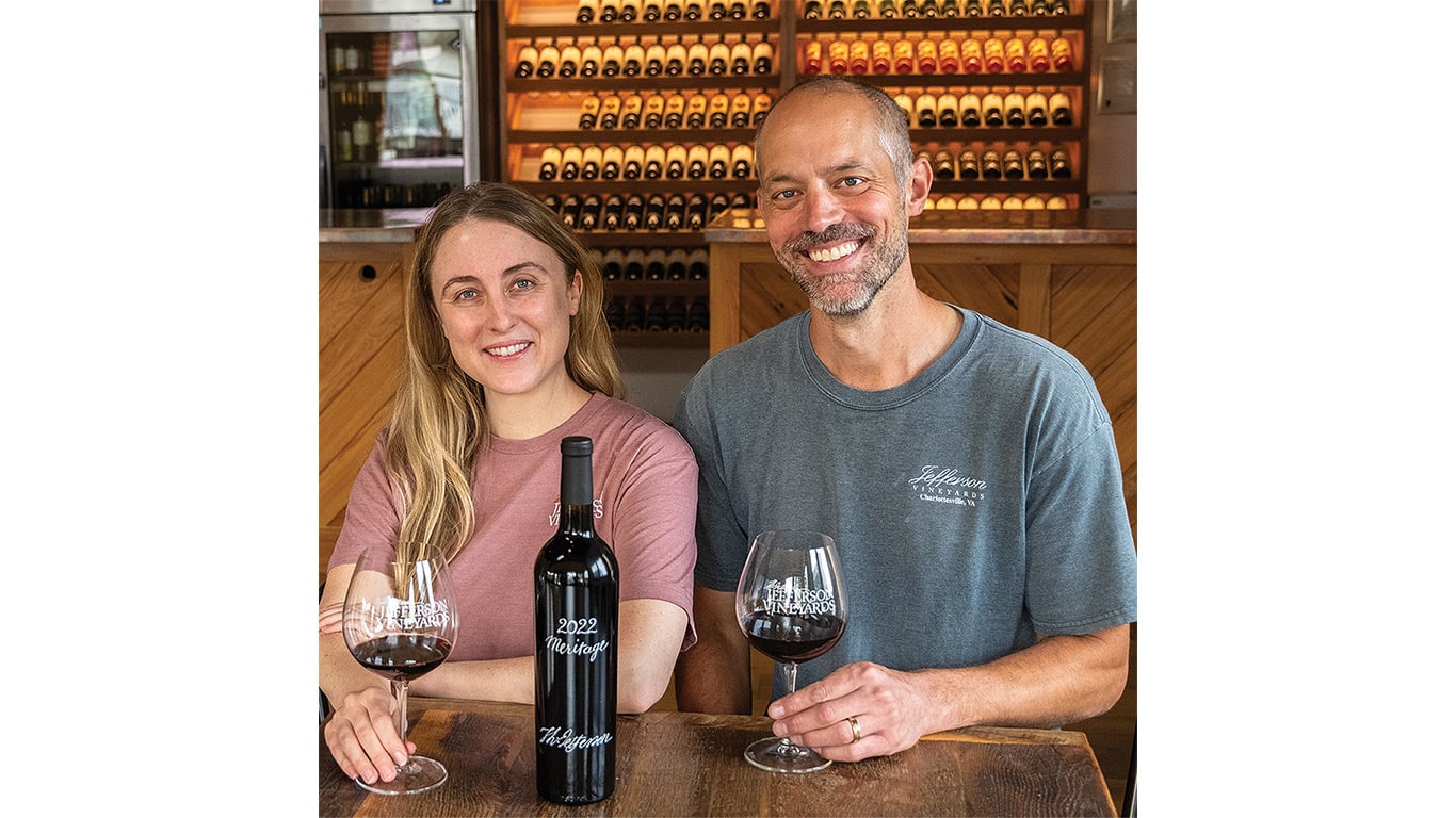 Two people seated at a table holding glasses of red wine with a wine bottle between them and shelves of bottles behind.