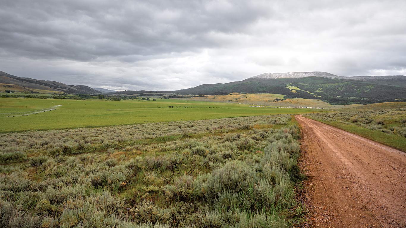 Dirt road runs through grassy valley toward distant hills under cloudy sky.
