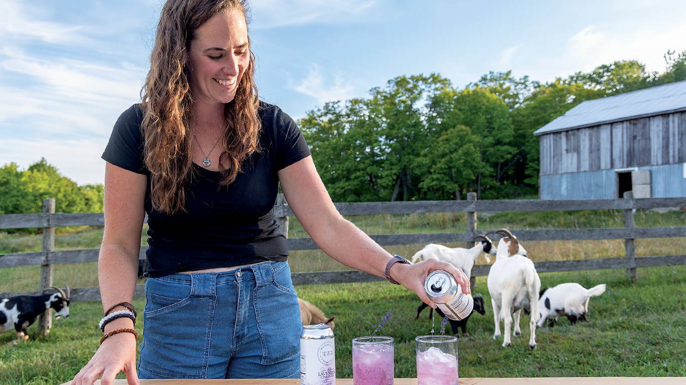 Person pouring a lavender drink outdoors with goats and a barn behind.