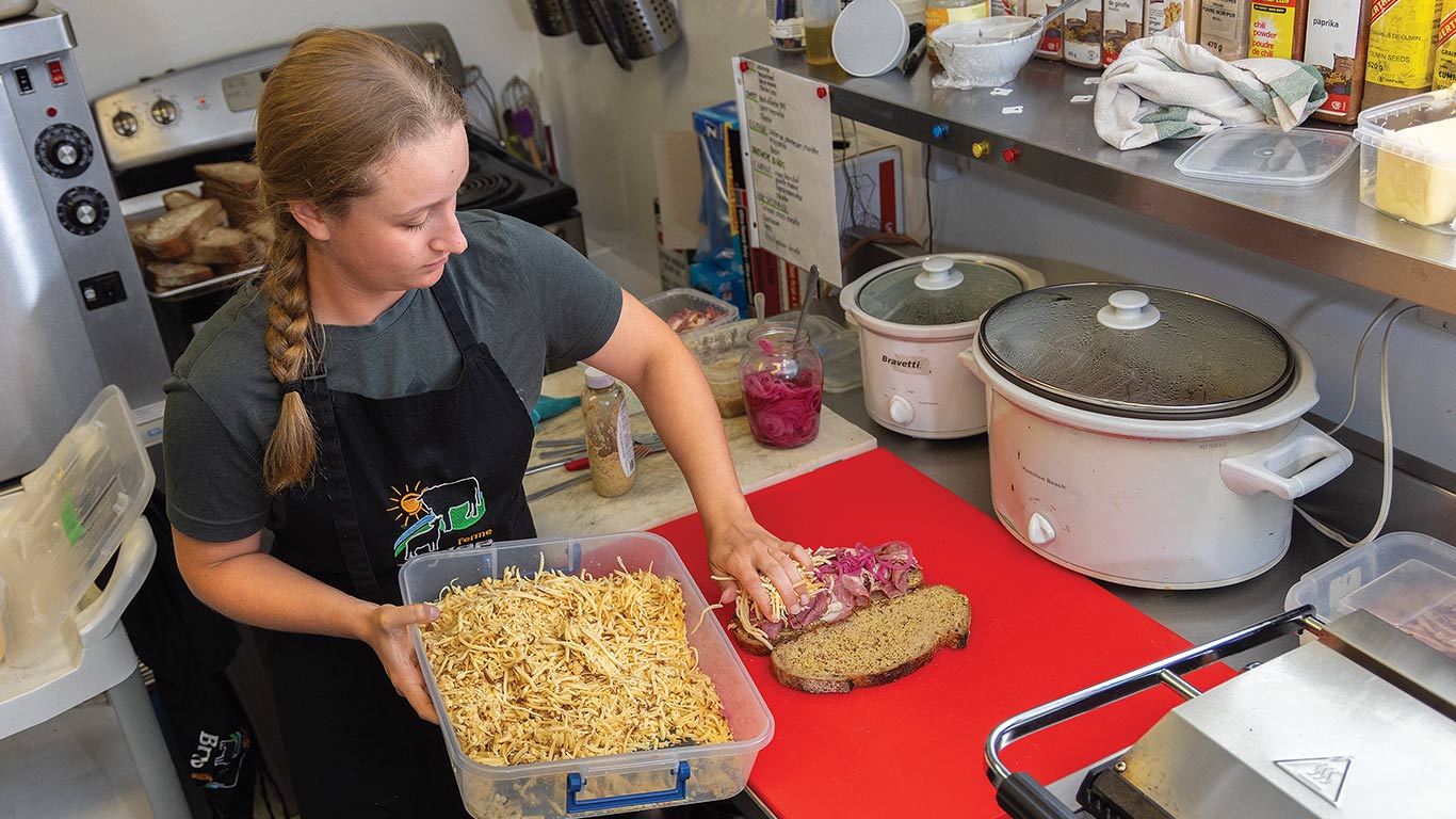 Person assembles sandwich in kitchen, adding shredded topping on bread beside slow cookers.