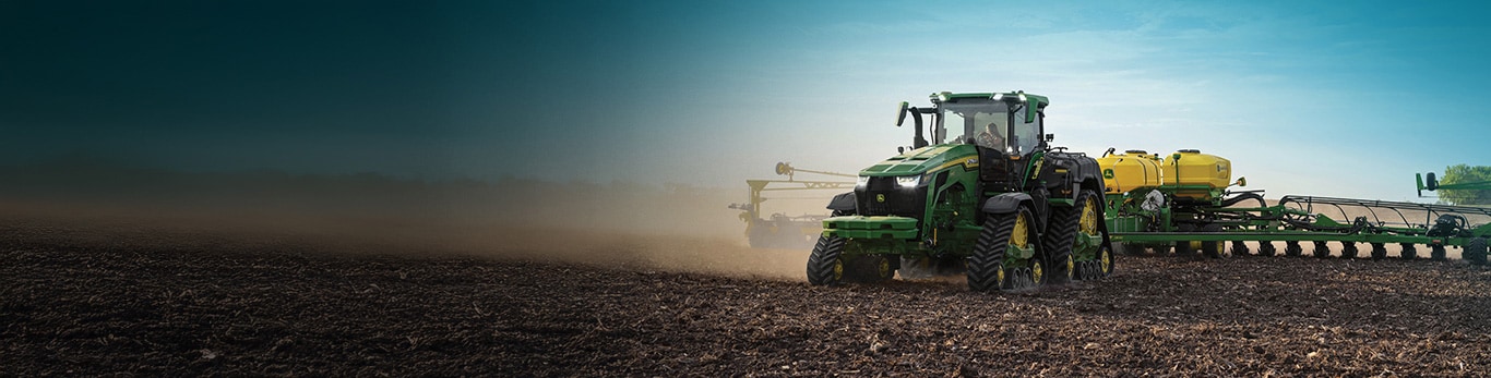Large tracked tractor planting crops in a wide field, with dust rising behind under a blue sky.